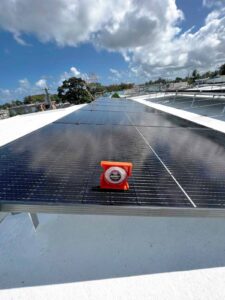 Un inclinómetro naranja colocado sobre un panel solar en un techo con múltiples paneles y cielo parcialmente nublado de fondo.