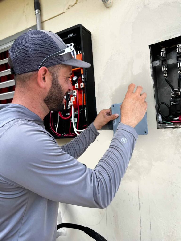 Un electricista sonriendo mientras instala o conecta componentes eléctricos en un sistema de baterías Tesla en una vivienda.